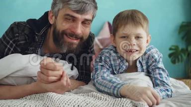 特写肖像两人成年父亲和可爱的小儿子躺在家里的床上微笑。 父爱